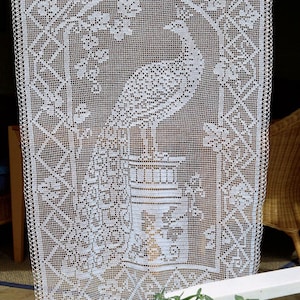 May include: A white, open-weave lace curtain featuring a detailed peacock design. The peacock is centered, with its tail fanned out. The curtain has a geometric border and floral accents, creating a delicate and ornate appearance.