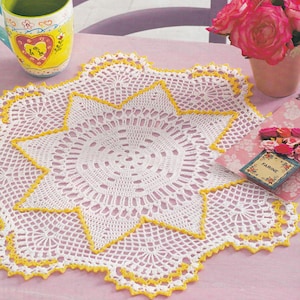 May include: White crochet doily with a yellow border, featuring a starburst design. The doily is on a pink table with a mug and flowers in the background.