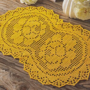 May include: A handmade, yellow crochet table runner with a detailed openwork design. The runner has a scalloped edge and is placed on a wooden surface. Text in the image reads: Häkelschrift und Zählmuster siehe Seite 26.