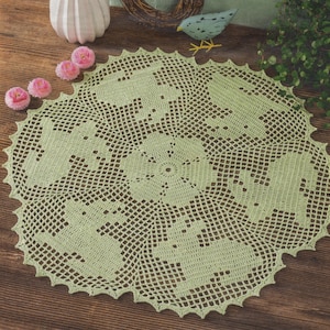 May include: A light green, crocheted doily with a scalloped edge. The doily features a repeating pattern of bunny rabbits and a central floral design. The handmade piece is displayed on a wooden surface, with decorative elements in the background.