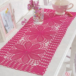 Könnte beinhalten: Ein rosa gehäkelter Tischläufer mit einem zarten Blumenmuster. Der Läufer liegt auf einem weißen Tisch mit einer rosa-weißen Teetasse und einer Glasvase mit rosa Blumen.