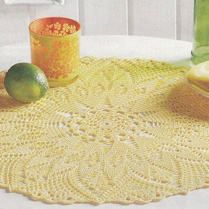 May include: A yellow crocheted doily with a delicate pattern sits on a white table with limes and a glass candle holder.