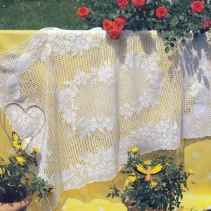 Puede incluir: Un mantel de ganchillo blanco con estampado floral drapeado sobre una superficie amarilla. El mantel presenta intrincados detalles de encaje y está rodeado de plantas en macetas con flores amarillas y rojas. Una decoración de corazón y mariposa de metal está en primer plano.