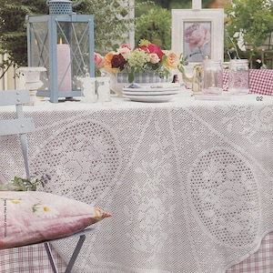 May include: A white crocheted tablecloth with a delicate floral pattern covers a table set for an outdoor meal. A blue lantern with a pink candle sits on the table, along with a vase of pink and red roses. Two clear glass jars with pink and white checkered lids are on the table, and a white picture frame with a floral print is leaning against a white easel.