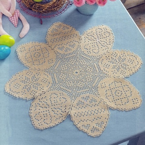 May include: A beige, flower-shaped doily with a detailed, openwork design. The doily is made of a crocheted material and has eight petal-like sections. The doily is placed on a light blue surface, with other decorative items in the background.