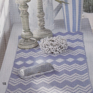 Può includere: Un runner da tavolo all'uncinetto con un motivo a chevron in tonalità di viola e bianco. Il runner è decorato con una scatola d'argento e un bouquet di fiori bianchi.