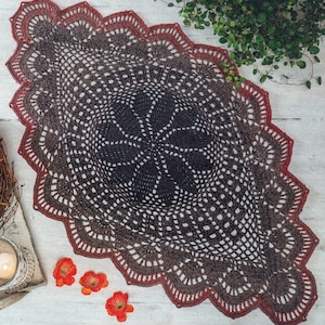 Könnte beinhalten: Handgefertigte, ovale Häkeldecke in Braun- und Dunkelrottönen. Das aufwendige Lochmuster-Design weist ein zentrales Blumenmuster und einen gewellten Rand auf. Die Decke wird auf einer hellen Holzoberfläche mit kleinen orangefarbenen Blumen und Grünzeug im Hintergrund präsentiert.