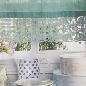 Può includere: Un'anfora bianca con pois blu si trova su una superficie bianca con un cumulo di piatti bianchi. Dietro l'anfora, una finestra con una tenda verde all'uncinetto con un bordo bianco. La tenda ha un bordo in pizzo all'uncinetto.