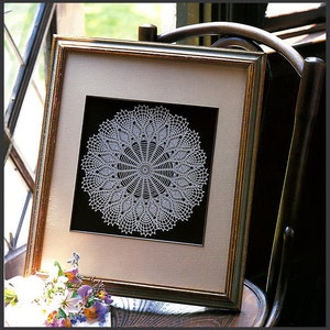 May include: A white crocheted doily framed in a gold frame with a black mat. The frame is sitting on a wooden chair.
