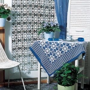 May include: A white crocheted curtain and a blue and white crocheted tablecloth are featured in this image. The tablecloth is on a small white table with a blue curtain in the background. Blue glass candle holders and a teacup and saucer are on the table.