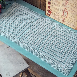 May include: A white, rectangular, crocheted doily with a geometric design, placed on a light blue surface. The doily has a detailed, openwork pattern and a scalloped edge. A vintage recipe card and pink roses are in the background.