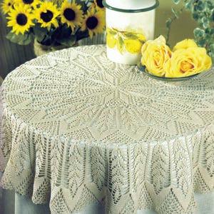 Op de afbeelding: Een beige, ronde, gehaakte tafelkleed met een gedetailleerd bladerpatroon. Het tafelkleed is over een ronde tafel gedrapeerd. Gele rozen en zonnebloemen zijn zichtbaar op de achtergrond, samen met een decoratieve pot met een citroenontwerp.