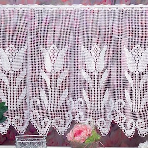 May include: White crocheted curtain with a floral tulip design. The curtain features a scalloped edge and is hung on a white rod. The intricate pattern is visible throughout the curtain, adding a delicate touch to the window treatment.