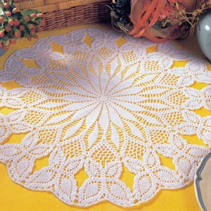May include: A white, crocheted doily with a detailed floral pattern. The circular doily features a central starburst design, surrounded by intricate openwork and a scalloped edge. The doily is displayed on a yellow surface, with other decorative items in the background.