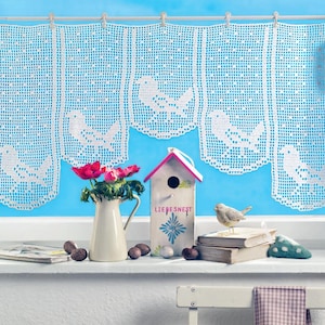 May include: White crocheted window curtains with bird designs hang in front of a blue sky. A white pitcher with red flowers, a birdhouse with the text "LIEBESNEST", and other decorative items sit on the windowsill.