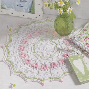 Könnte beinhalten: Handgefertigte, weiße Häkel-Tischdecke mit floralem Design. Sie hat rosa und grüne Akzente und einen gewellten Rand. Die runde Tischdecke scheint aus Baumwollgarn gefertigt zu sein. Ein dekorativer Artikel für einen Tisch oder ein Regal.