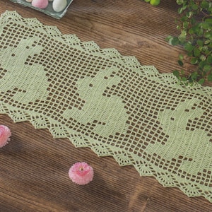 May include: A pale green, crocheted table runner featuring a bunny rabbit pattern. The runner is rectangular with a scalloped edge. Pink flowers and decorative eggs are scattered on a wooden surface.