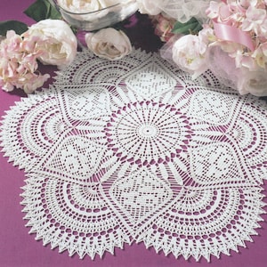 May include: A white, intricately crocheted doily with a detailed geometric pattern. The doily is oval-shaped with a scalloped edge. The background includes pink and white flowers, suggesting a vintage or handmade aesthetic. The doily is likely used for decorative purposes.