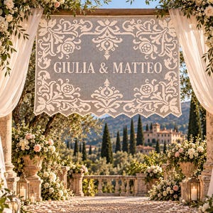 May include: A rectangular white lace banner featuring the names GIULIA & MATTEO in bold capital letters, surrounded by an intricate floral pattern. The banner is suspended between two stone pillars adorned with white roses and green foliage.