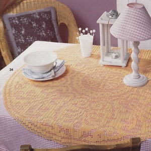 May include: A crocheted table runner with a delicate floral design in a light yellow color with pink accents. The runner is laid on a white tablecloth with a pink and white checkered pattern. A white bowl, a white lantern, and a white lamp with a pink and white checkered shade are on the table.