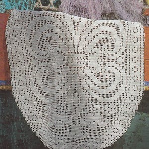 Puede incluir: Un tapete blanco, intrincadamente tejido a ganchillo con un diseño floral detallado. El tapete está drapeado sobre una superficie oscura, mostrando su delicado patrón calado. Los bordes están festoneados, agregando un toque decorativo a este textil hecho a mano.