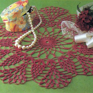 Può includere: Un centrino all'uncinetto bordeaux con un motivo floreale dettagliato. Una collana di perle e una scatola floreale sono sul centrino. È presente anche un piccolo bouquet di fiori rossi con un nastro bianco. Il centrino è su una superficie verde.