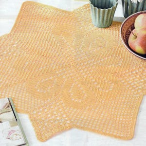 Puede incluir: Un tapete de ganchillo en forma de estrella, en un tono amarillo suave. El intrincado diseño calado es visible en toda la pieza. El tapete está colocado sobre una superficie blanca, con un cuenco de manzanas y un pequeño jarrón verde cerca. También hay una revista.