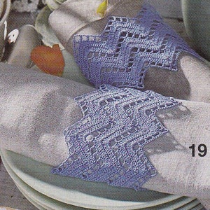 Può includere: Un primo piano di un tovagliolo di lino grigio chiaro con un bordo di pizzo all'uncinetto blu. Il tovagliolo è piegato e posizionato su un piatto bianco. Il numero "19" è stampato sul tovagliolo.