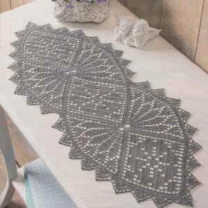 May include: A gray, handmade crochet table runner with a detailed, openwork design. The runner has a scalloped edge and a geometric pattern. The runner is displayed on a white surface.