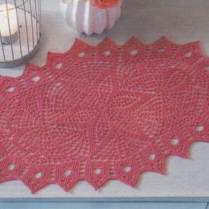 May include: A handmade, oval-shaped, coral-colored crochet doily. The doily features a detailed, openwork design with a scalloped edge. The doily is placed on a light-colored surface, with a candle in a metal cage and a white vase in the background.