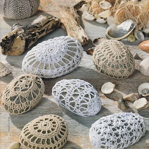 Könnte beinhalten: Sechs Häkelüberzüge für Steine in Weiß und Beige. Die Überzüge sind mit einem zarten Spitzenmuster gearbeitet und werden auf einer Holzoberfläche mit Muscheln präsentiert.