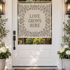 May include: A square, off-white crochet wall hanging featuring a dark brown floral border of roses and leaves surrounding the centered text "LOVE GROWS HERE" in a bold, block font, suspended from a thin metal rod across a glass door panel.