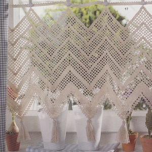 Può includere: Un tendaggio per finestra all'uncinetto con un motivo a chevron e frange. Il tendaggio è beige e pende da un'asta bianca. Il tendaggio è davanti a una finestra con cornice bianca e vista su piante verdi.