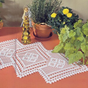 May include: A white, crocheted table runner with a geometric pattern and scalloped edges. A decorative glass bottle filled with oil sits to the left, and potted plants with yellow flowers and green foliage are in the background.