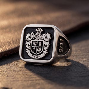 Puede incluir: Un anillo de sello de plata con una cara cuadrada y un fondo negro hundido. El centro muestra un escudo heráldico detallado con el nombre BEAN en una cinta debajo. El lateral del anillo está grabado con el texto 9/11 en letra negrita.