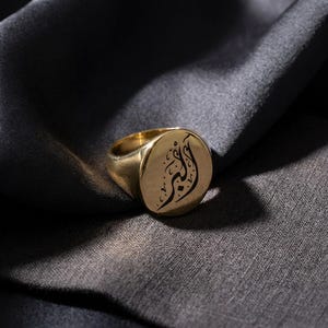 May include: A gold signet ring with an oval face. The ring features black calligraphy on the face. The ring is set against a dark gray fabric background. The ring is a classic piece of jewelry.
