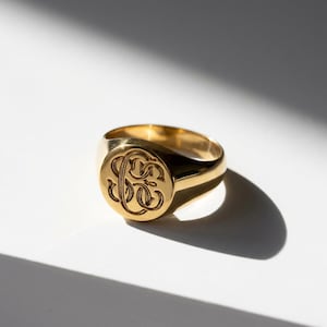 Könnte beinhalten: Ein goldener Siegelring mit eingraviertem Monogramm. Der Ring hat eine polierte Oberfläche und eine abgerundete Oberfläche mit einem eingravierten Design. Der Ring ist vor einem weißen Hintergrund mit einem Schatten platziert.