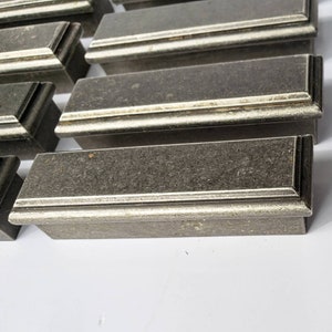 May include: A set of eight silver-toned drawer pulls with a rectangular shape and a raised edge. The pulls have a brushed finish and a subtle texture.