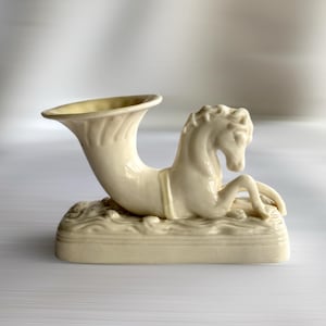 May include: Cream-colored ceramic vase with a horse design. The vase features a horn-like opening and a horse figure emerging from a base with wave-like details. The horse is in a reclining position, adding a decorative touch to the piece.