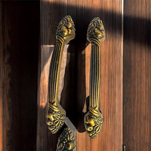 May include: Ornate gold-colored door handles with a detailed floral design. The handles are mounted on a dark brown wooden door, with vertical wood grain visible. The handles have a vertical ribbed design.