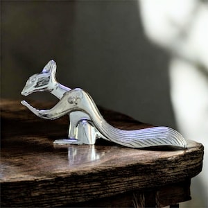 May include: A chrome-plated squirrel figurine with a detailed design. The squirrel is posed in a dynamic stance, with its tail curved and its head raised. The figurine is set on a wooden surface.