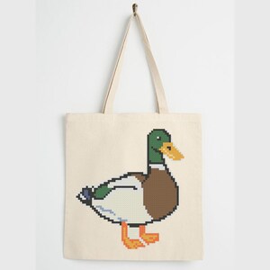 Puede incluir: Un bolso de tela beige con un gráfico de pixel art de un pato real. El pato tiene la cabeza verde, el pico amarillo, el pecho marrón, plumas blancas y patas naranjas. El bolso cuenta con dos asas largas para el hombro y forma rectangular.