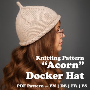 Può includere: Un cappello Docker "Acorn" beige lavorato a maglia con una corona arrotondata e una piccola sommità a punta. Il cappello è mostrato su una persona con lunghi capelli castani ondulati. L'immagine include il testo "Knitting Pattern", "Acorn", "Docker Hat" e "PDF Pattern - EN | DE | FR | ES".