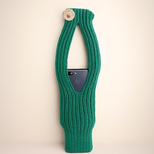 May include: A green knitted phone holder with a wooden button closure. The holder is designed to cradle a smartphone, with an opening for the screen and a loop for carrying. The holder is made of a thick, textured yarn.