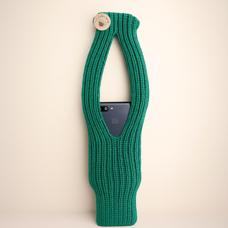 May include: A green knitted phone holder with a wooden button closure. The holder is designed to cradle a smartphone, with an opening for the screen and a loop for carrying. The holder is made of a thick, textured yarn.