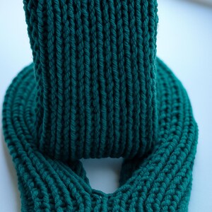 May include: A close-up of a teal-colored knitted scarf. The scarf is made of thick yarn and has a ribbed texture. The scarf is folded over itself, creating a loop. The background is a light, solid color.