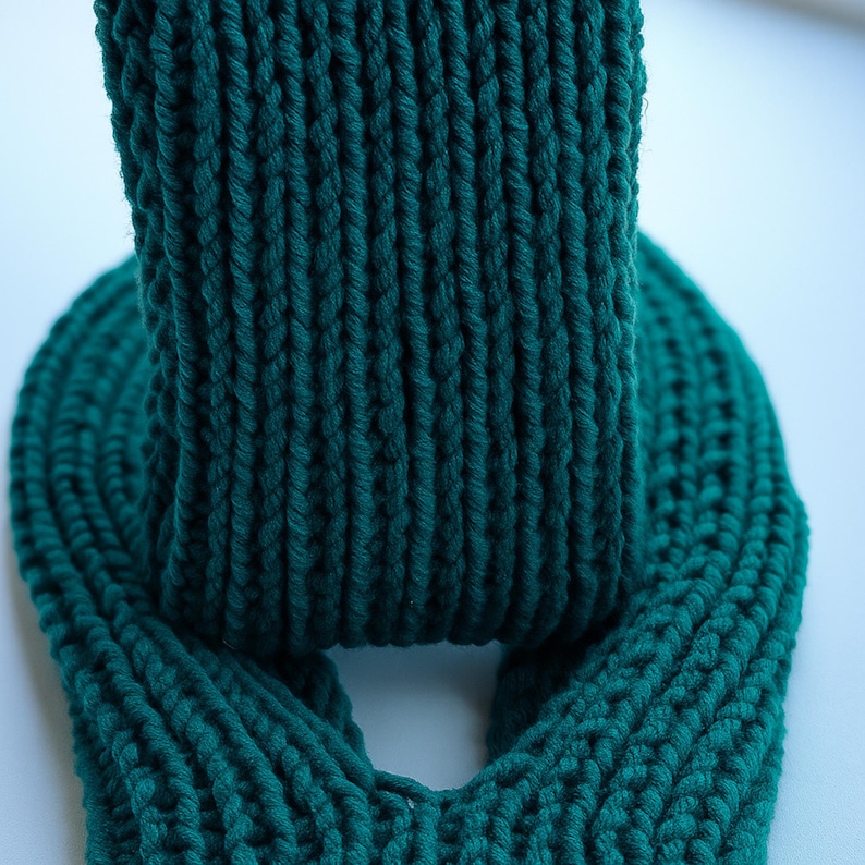 May include: A close-up of a teal-colored knitted scarf. The scarf is made of thick yarn and has a ribbed texture. The scarf is folded over itself, creating a loop. The background is a light, solid color.
