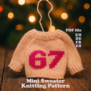 Könnte beinhalten: Ein kleiner, beigefarbener Strickpullover mit der Zahl 67 in Pink. Er hat eine Schlaufe zum Aufhängen und den Text "Mini Sweater Knitting Pattern" unten. Der Hintergrund ist verschwommen mit Bokeh-Lichtern.