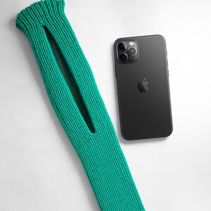 May include: A teal green knitted sleeve with a large opening, next to a black smartphone with a triple-lens camera. The sleeve is made of a thick, textured yarn and appears to be a protective cover for a device.