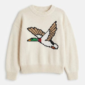 May include: Cream-colored knit sweater featuring a pixelated design of a flying duck. The duck has brown, white, black, green, red, and yellow details. The sweater has a ribbed crew neck, cuffs, and hem.